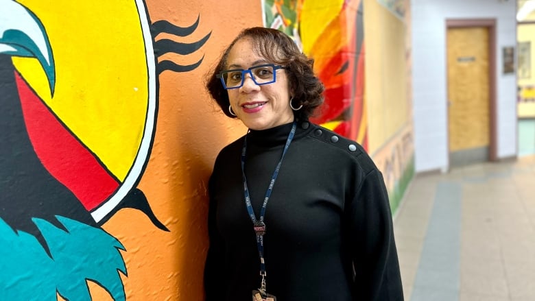 A woman with glasses stands in front of a colourfully painted wall.