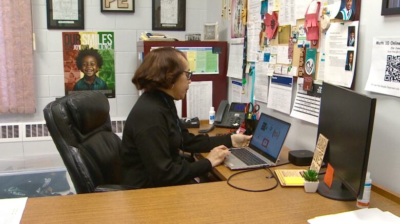 A woman sits at a desk with a computer and a number of papers on the wall.