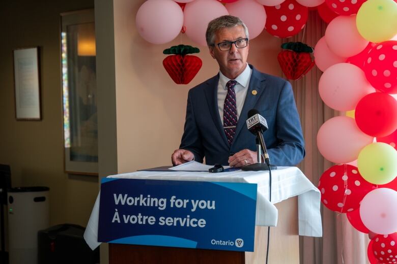 A person wearing a suit is seen standing at a podium indoors and speaking into a microphone. Behind them is an archway of pink, yellow, and red and white polka dot balloons.