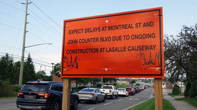 A big orange sign warns drivers to expect delays. Behind it is a long line of cars waiting at a stoplight.