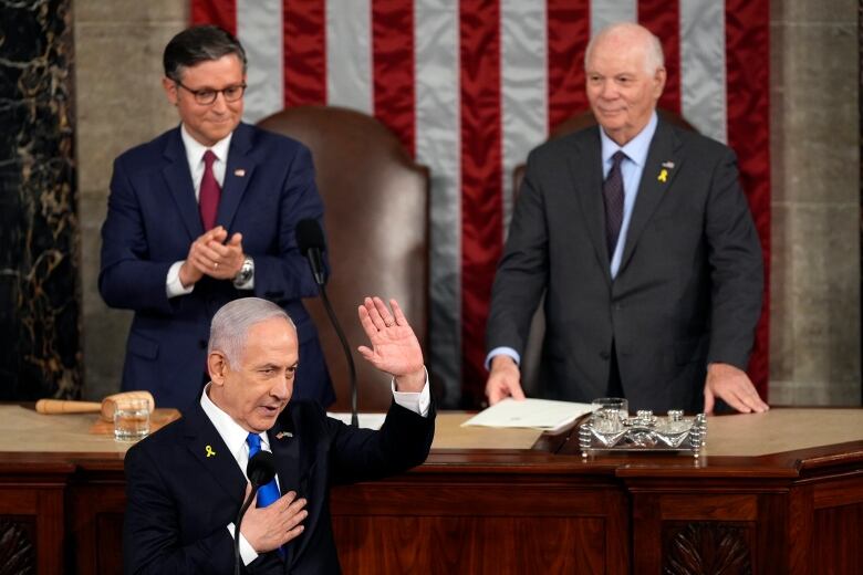 A person waves and holds their hand to their chest as other behind them clap and stand.