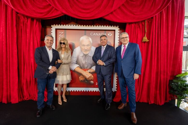 Rick Mercer, Lynne St. David Jewison,  Charles Sousa, and Doug Ettinger stand on stage in front of a red curtain unveilling the new stamp with a photo of Norman Jewison,