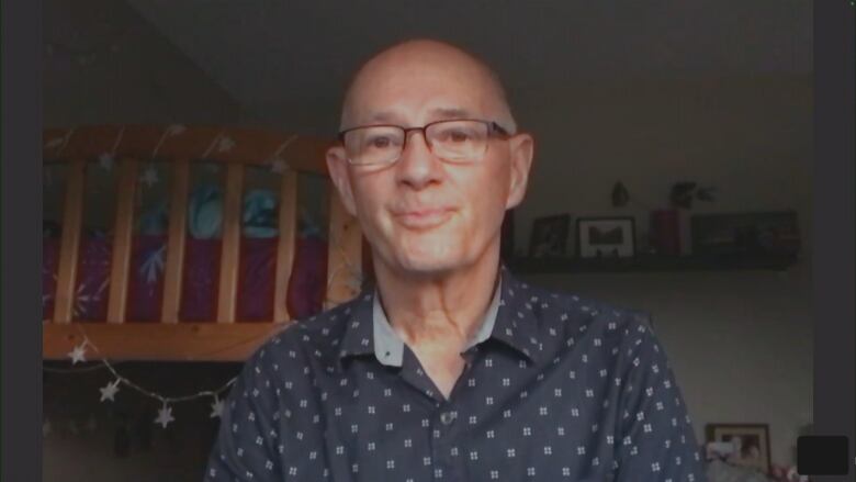 A bald white man wearing a navy blue button-up shirt is sitting in a bedroom with a bunkbed behind him.