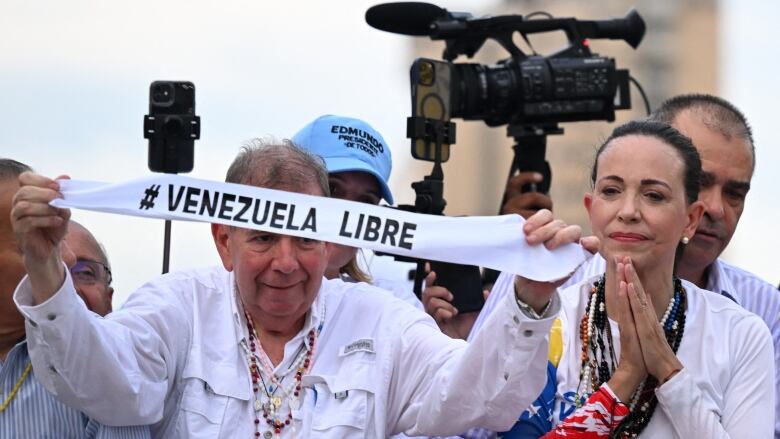 An older cleanshaven man holds a banner that says 'Venezuela Libre' as a woman with her hair pulled back looks on to his left.