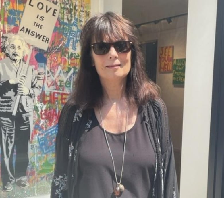 A woman wearing mostly black with sunglasses and a long necklace smiles for a photo. A graffiti art mural is in the background featuring an image of Albert Einstein holding a sign that reads 