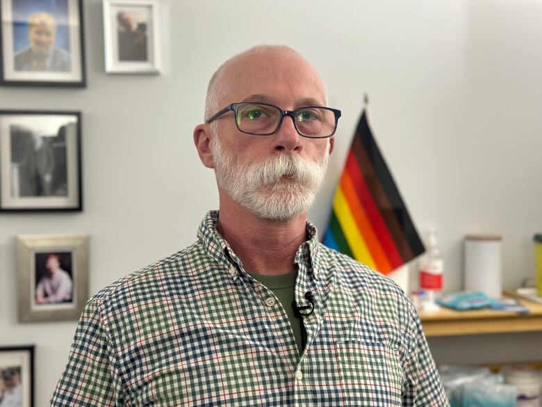 a man wearing glasses and a button up shirt looks into the canera. A pride flag and portrait are in the background. 