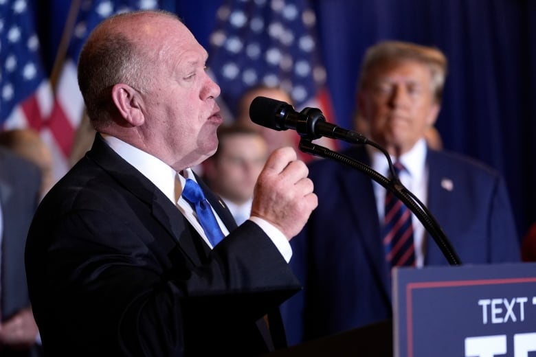 A man in a suit gestures while speaking at a lectern. Other men, and some American flags, are visible in the blurry background. 