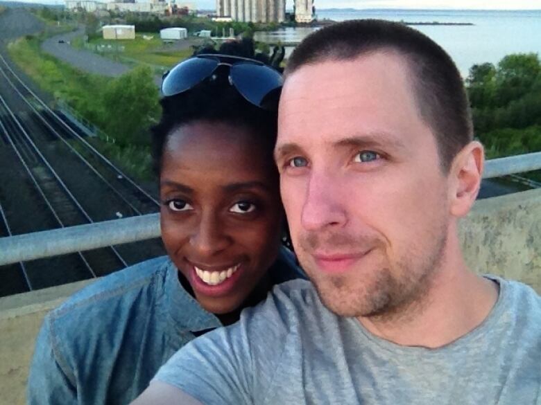 A man and woman, standing close together, smile for the camera. Railroad tracks and a body of water are in the distance. 