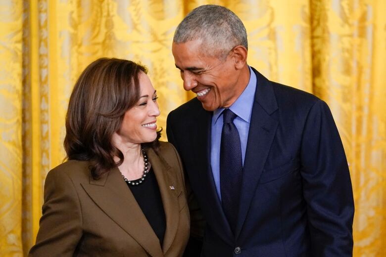 A man and a woman, both darkskinned, smile while standing next to each other wearing blazers.