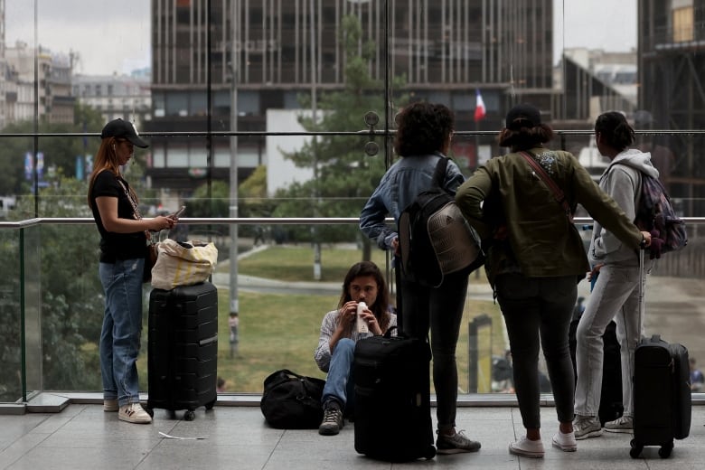 People are shown seated on the ground and standing with luggage inside a building.