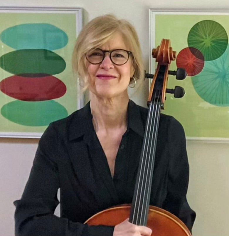 A woman with blonde hair and dark glasses, smiles softly for the camera as she sits holding a cello.