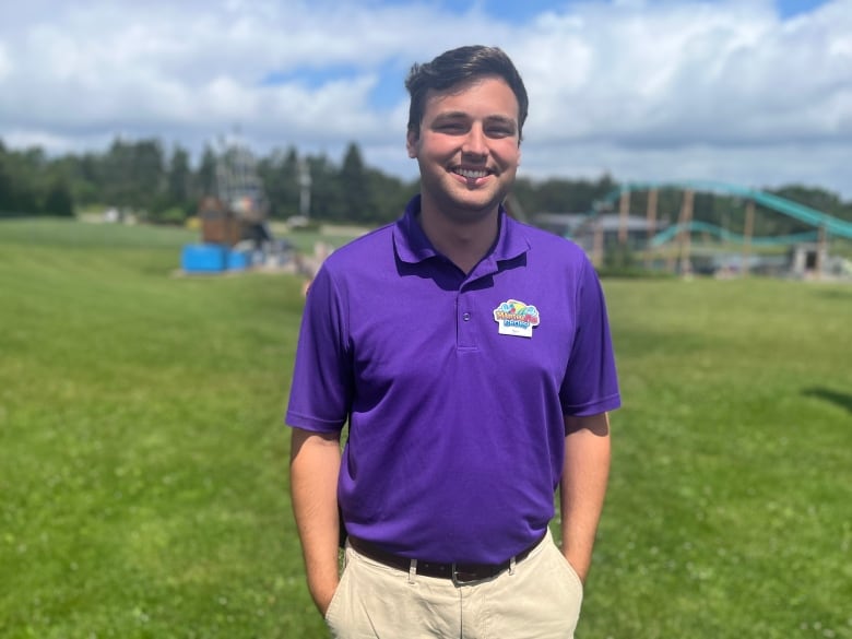 A man in a bright purple t-shirt stands inside a water theme park with a rollercoaster behind him.