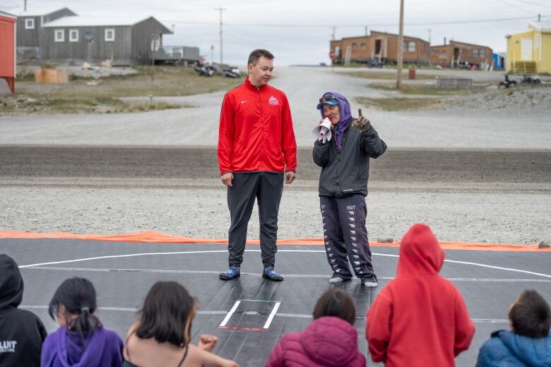 two men stand on a black wrestling mat, one holds a microphone. children watch them.