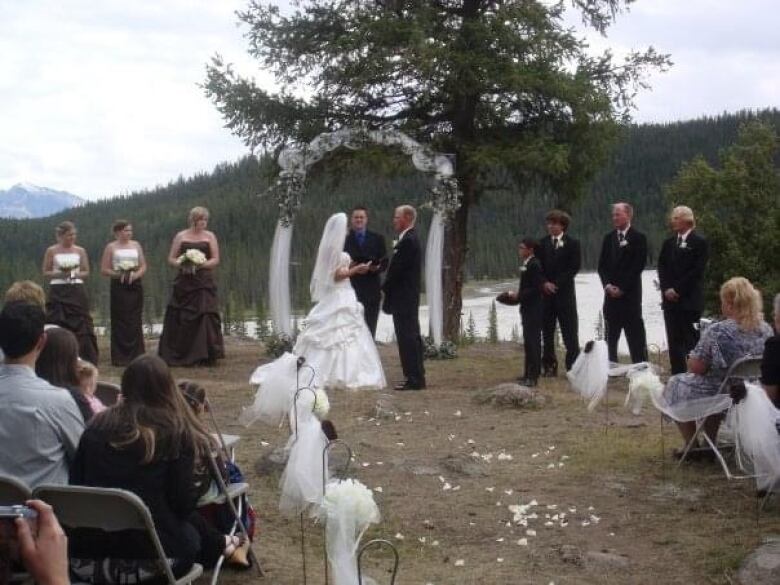 A man and woman are giving wedding vows in front of a pastor in an outdoor wedding in front of a river. 