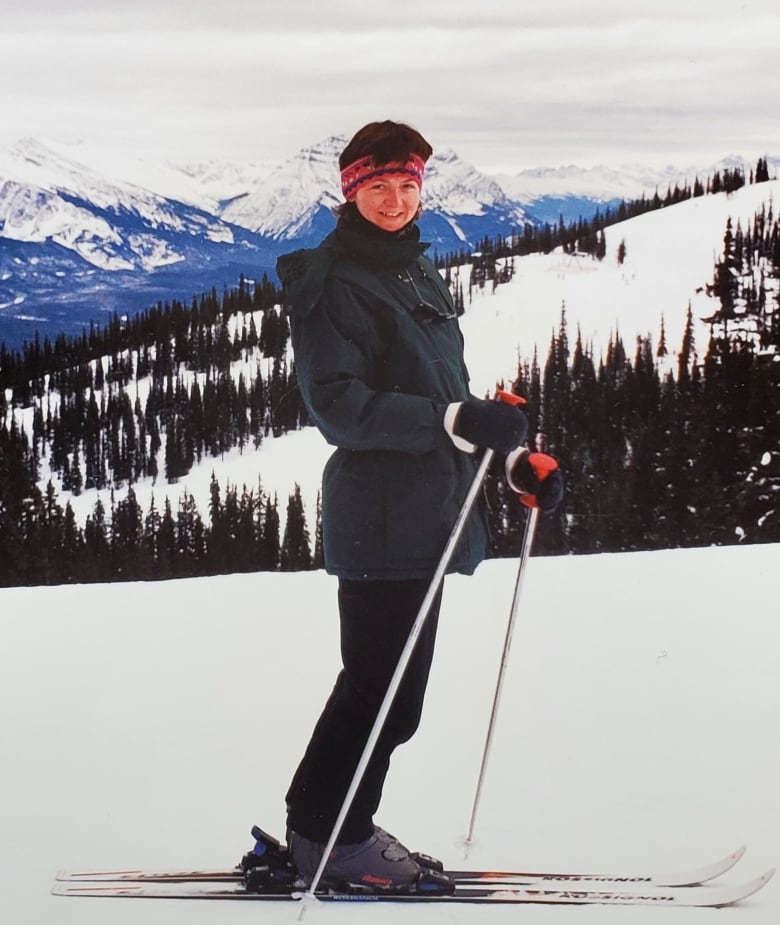 Woman in skit suit on skis poses for camera on a ski run. Evergreen trees and other ski runs can be seen in the near background. Other mountains can be seen in the distant background.