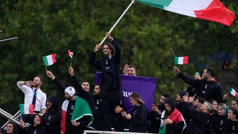 A man waves an Italian flag while standing on top of a boat during a parade along a river.