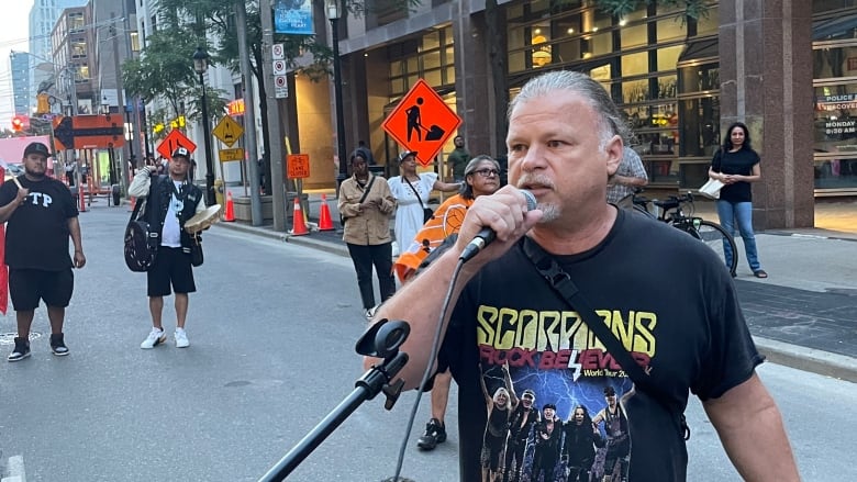 A middle aged man with grey hair and a goatee speaks into a microphone at a rally on a city street.