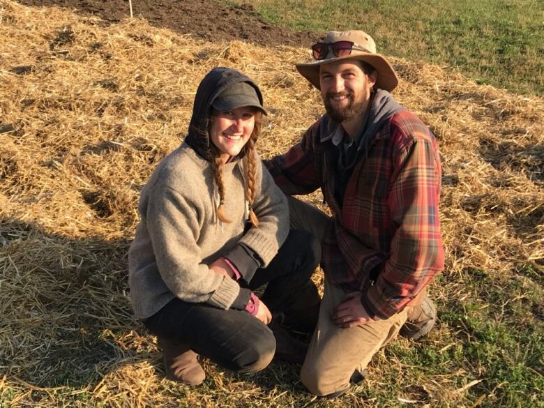 A woman and a man pose on farmland surrounded by hay. 