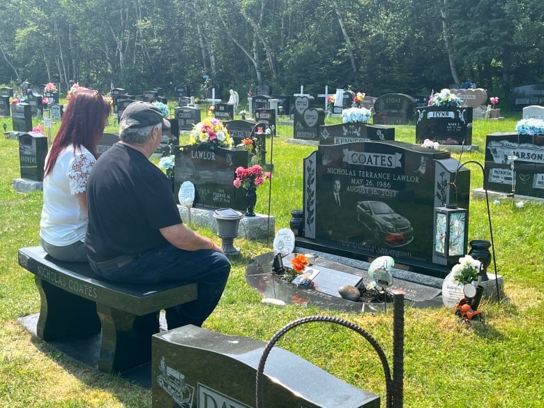 A woman and a man sitting on bench in front of a grave of young man. 