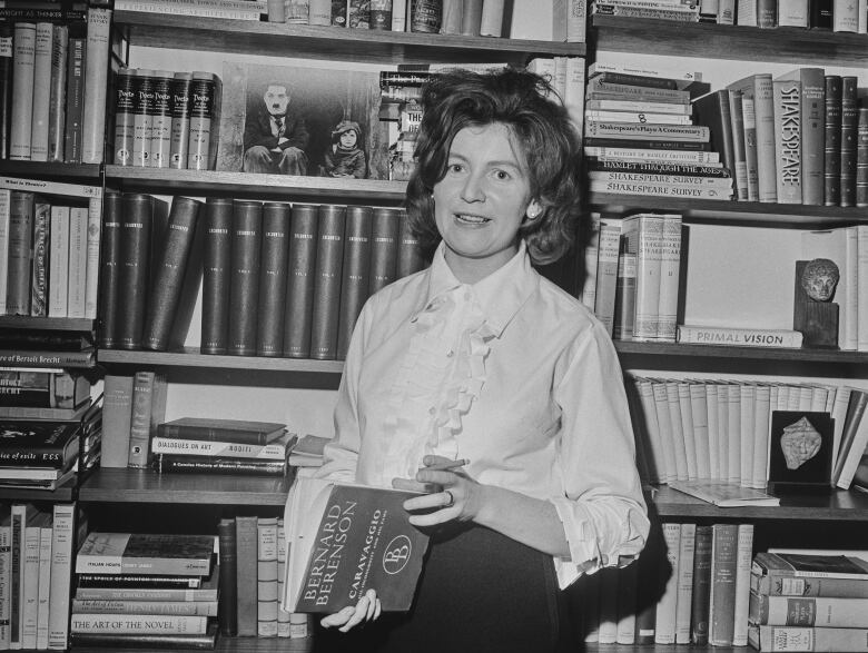 In this black and white photograph, a person holding a book and a cigarette poses in front of a book shelf.