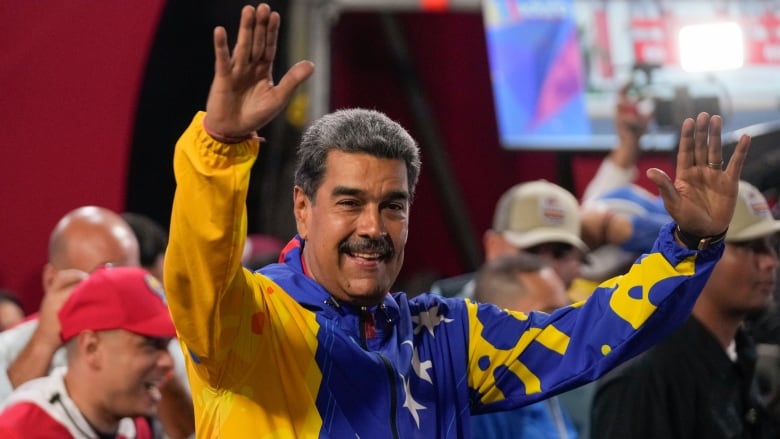 A man in a purple and yellow track jacket smiles and raises both his arms at a crowd.