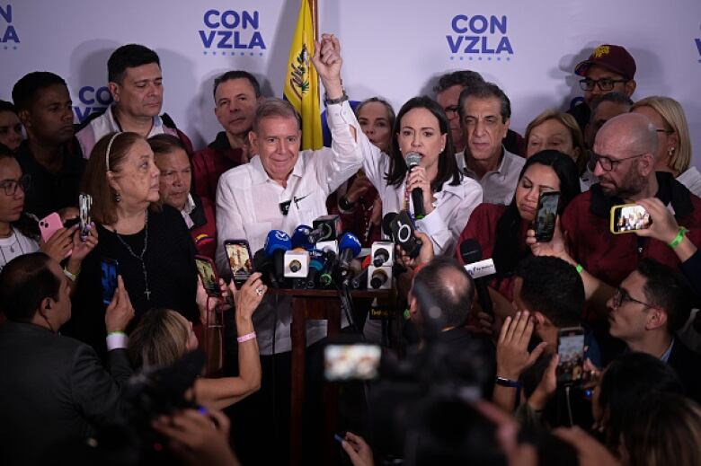 A man and woman, both dressed in white shirts, are surrounded by reporters with microphone and cameras. The woman in white speaks into a microphone.