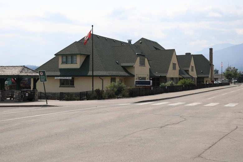 A long beige building with a steeply sloped greenish roof is shown from the street. A flagpole flies the Canadian flag in front.