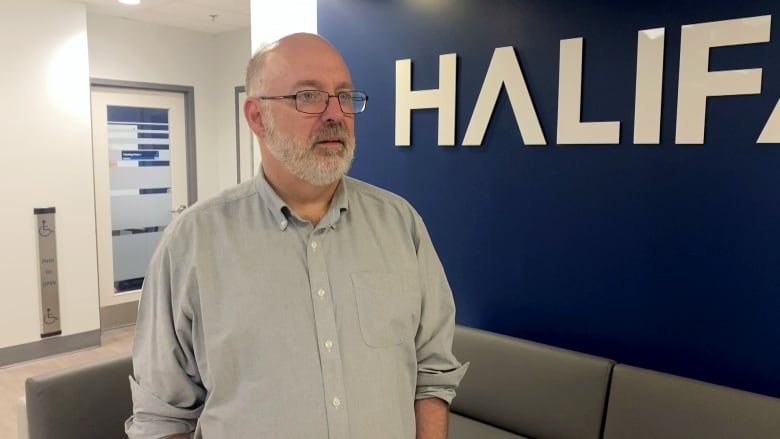 A white man with glasses and a white beard stands in front of a blue wall with the word HALIFAX in capital letters behind him