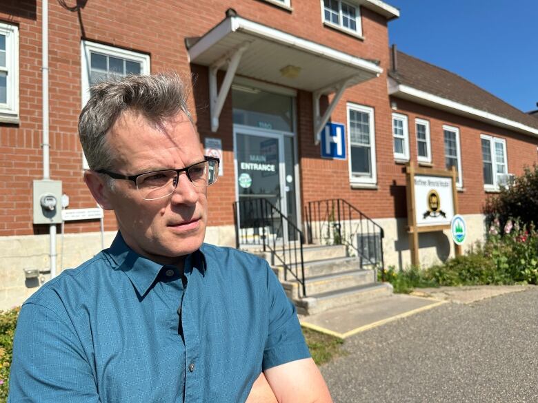 A man with short grey hair and glassess wearing a blue shirt stands in front of a red brick building with the universersal sign for a hospital, a white H on a blue background.