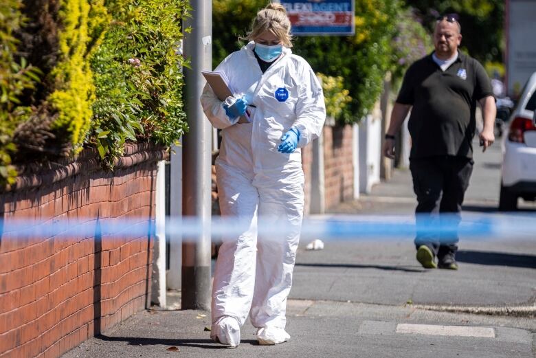 A woman wearing a protective suit and holding a clipboard walks along a sidewalk. There is police tape in the foreground. 