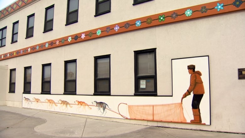 A cream-coloured building with a mural of a man driving a dogsled team