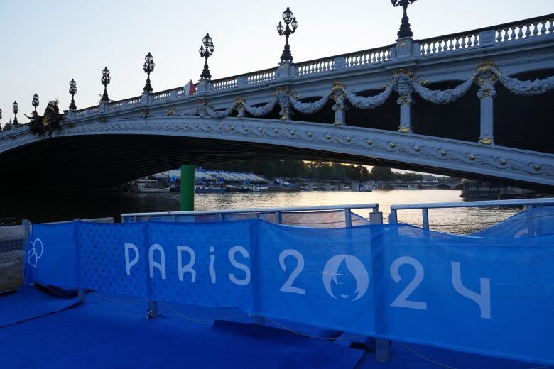 A bridge is shown spanning a river. Beneath it is a sign for the Paris 2024 Games.