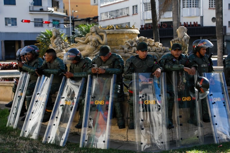 Police lean their backs against some statuary. Their riot shields are down and some of their helmets are off. A couple look at their smartphone.