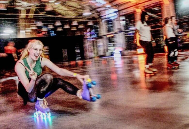 A blurry image of roller skaters in an indoor rink. One woman is on her knees and throwing the peace sign at the camera.