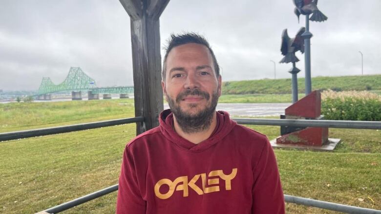 A man with brown facial hair and wearing a red sweater stands outside, a bridge and grass in the background.