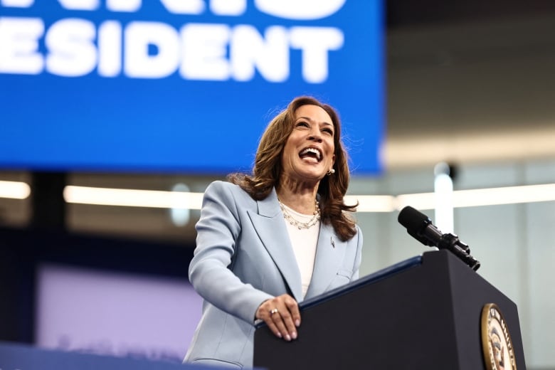 Kamala Harris, wearing a light blue blazer, smiles with a big, open-mouthed grin while standing at a podium.