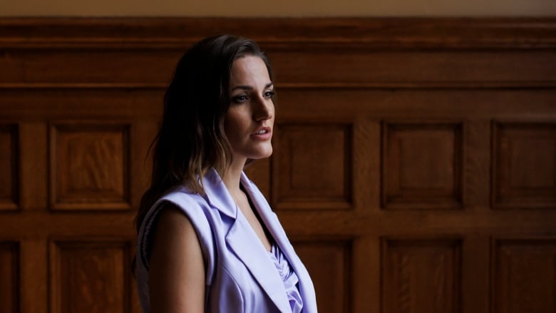 A woman in a lilac vets stands in front of a wodden panelled wall.