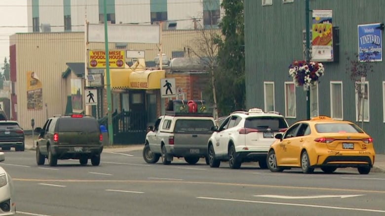 Vehicles including two taxis are parked by the side of a road in a city.