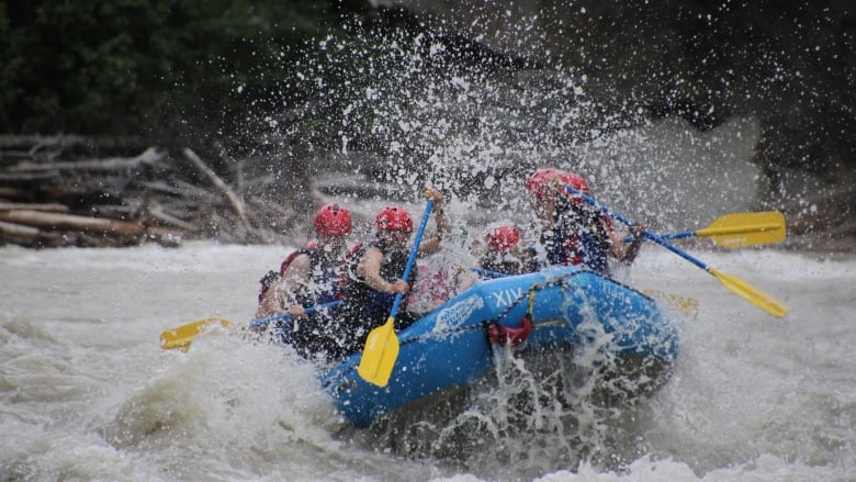 People wearing red helmets paddle through turbulent whitewater in a blue inflatable raft.