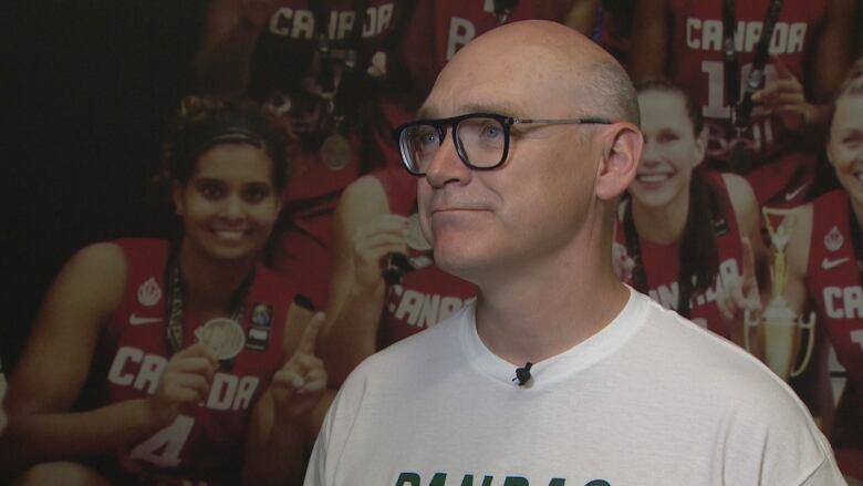 A bald white man with thick glasses is wearing a white t-shirt, while standing in front of a mural of women basketball players holding up medals.