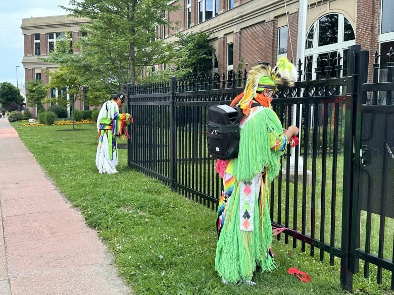 Two people dressed in colourful clothing stand at a short distance from each other, tying red cotton to a black fence.