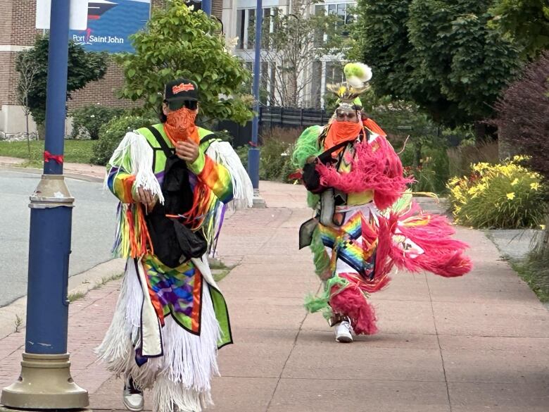 Two people dressed in colourful, ankle-length garments run down a sidewalk.