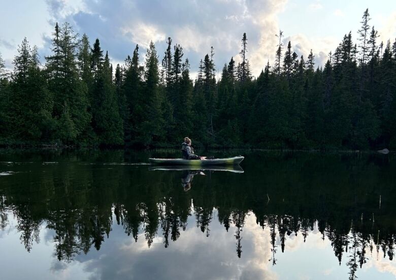 A person rests in a kayak floating on a still lake lined by trees.