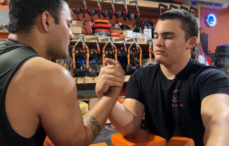 Two guys look serious as they link hands and prepare to arm wrestle. Their elbows are on orange blocks. 