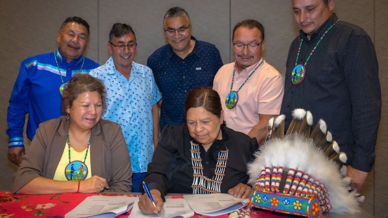 Several people stand or sit near a woman who signs a paper with a pen