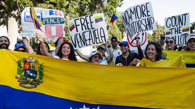 A group of people hold signs and stand before a blue, red and yellow flag with stars. 