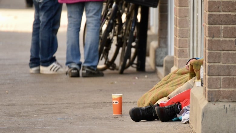 Feet of homeless person sticking out of doorway and coffee cup on the ground nearby. The legs of two other people are in the distance next to bicycles.