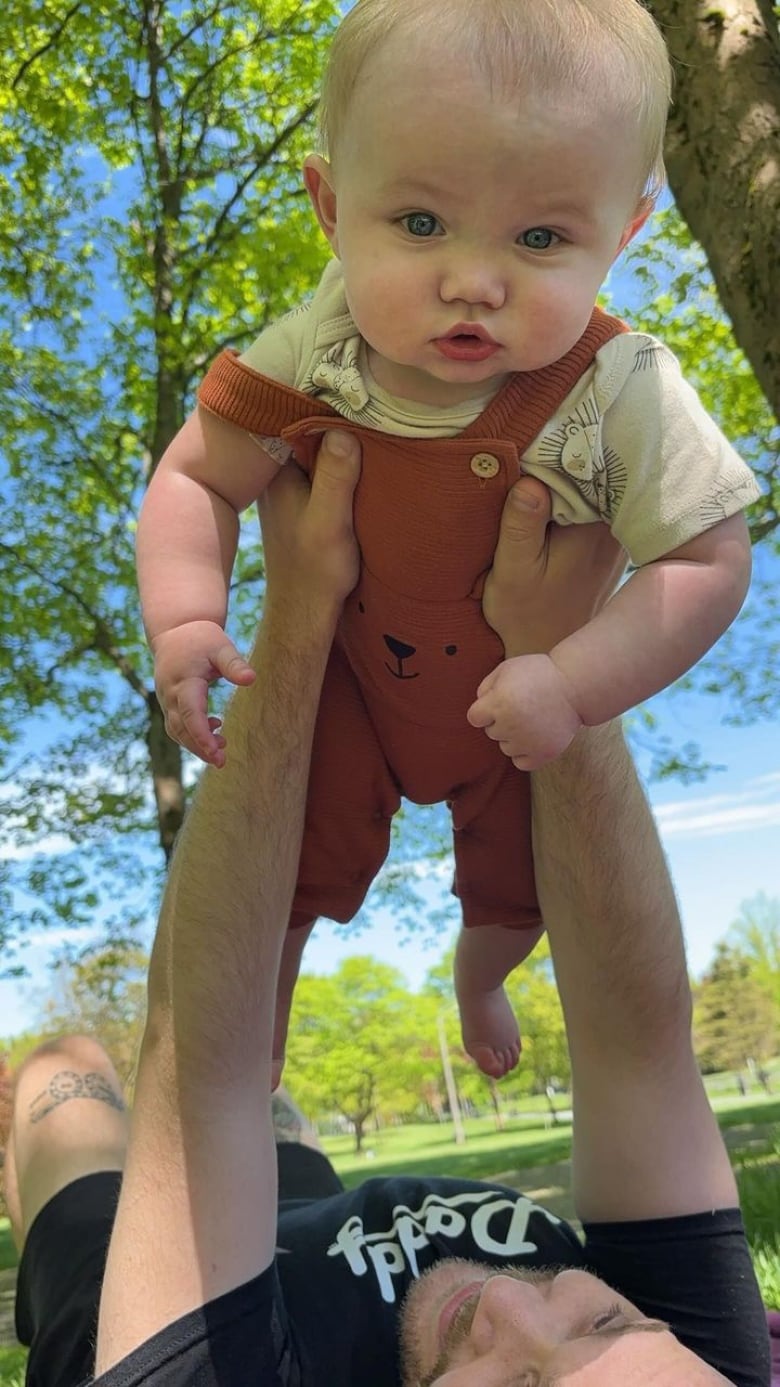 A man lying on his back in a park holds a baby in the air.