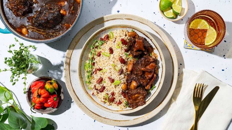 A bowl of oxtail stew with butter beans with a side of rice and peas, on a countertop with a Dutch oven in the background.