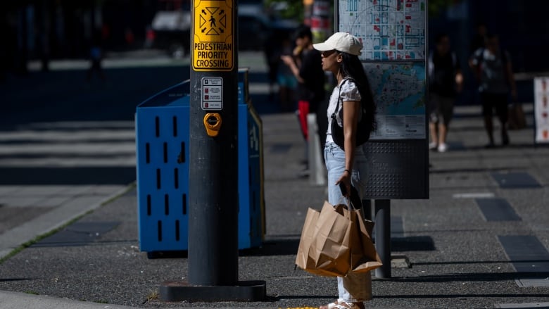 A lady in a cap carrying some paper bags and waiting to cross the road at a city street. 